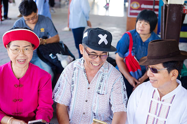 Three happy mature thai senior people are sitting close together on a bench on platform in station of Lampang. Lampang is in northern Thailand. All three are wearing headwear. One man and the woman are wearing eyewear and other man is wearing sunglasses. They are communicating and holding mobile phones. Man in center is smiling and looking at mobile phone. Woman is smiling and looking into the camera. Man at right side is looking into direction of woman and is smiling too