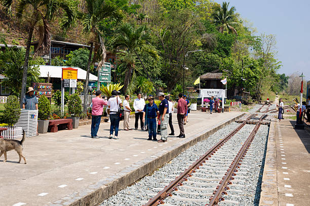 View along tracks at railway station Khun Tan in northern Thailand. Old station is located in Lamphun province. Close to station is a tunnel through mountains. Thai people are on platform and are standing in groups. Some are talking and some are taking photos. In right area is deadlock part of railway at small intersection of tracks. At far right area train is standing and partly visible. Khun Tan station is travel destination as part of thai railway history