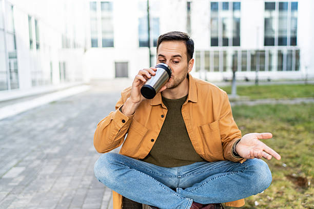 Young adult man sitting with crossed legs, drinking from a reusable coffee cup in a modern city park, enjoying a peaceful moment outdoors