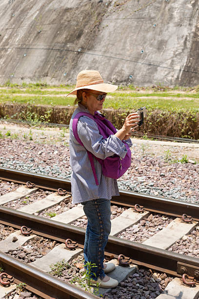 Mature thai woman with hat is taking photos with mobile phone while standing on tracks between station and tunnel Khun Tan in north Thailand. She has mobile in a telescope frame and stick and is preparing mobile for taking photo. She is standing with back to tunnel and front towards station both out of frame and sight. Station and tunnel are tourist attraction in Lamphun province