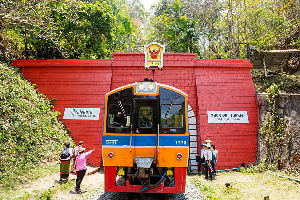 Front view of train at and inside of red colored tunnel Khun Tan. Tunnel is close to sttaion Khun Tan and part of travel and tourist attraction in Lamphun province. Station offers also access to Doi Khun Tan national park. A few thai people are at both sides of the train. Above train at tunnel is emblem of winged thai king Garuda which is national symbol of Thailand.