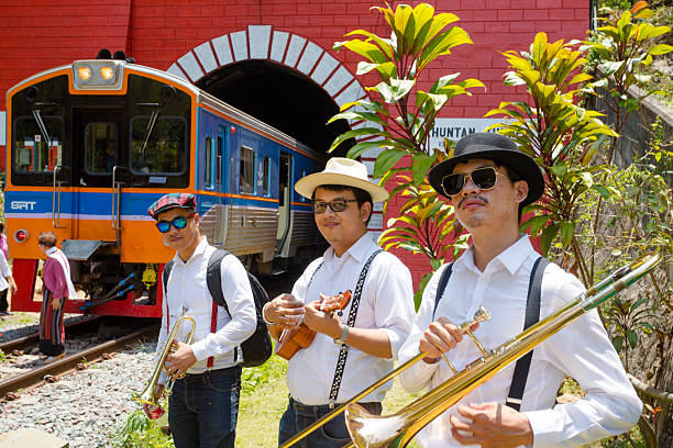 Three thai jazz musicians in front of train and tunnel Khun Tan near station Khun Tan. Men are standing aside tracks. The train is standing mostly inside of the tunnel. Some people and a senior thai women are at train. Tunnel and train are tourist and travel attraction in Lamphun province. Same time station offers access and entrance to Doi Khun Tan national park. Musicians are wearing headwear and eywear or sunglasses
