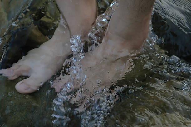Close-up of bare feet immersed in a shallow stream, with clear water flowing gently over the skin and stones beneath. The image evokes a sense of calm, grounding, and natural healing — ideal for concepts related to wellness, freedom, connection to nature, and outdoor relaxation.