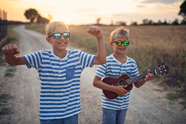 Two young boys stroll down a dusty rural path, bathed in the warm hues of the setting sun. Both twins are dressed in striped shirts and shorts. One of the boys carries a small guitar, suggesting a love for music or a playful adventure. The surrounding landscape features open fields. The golden light of sunset adds depth and warmth.\nShot with Nikon D850