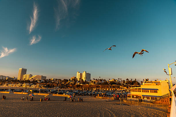 Bustling beach scene with a backdrop of a densely populated city. The image shows a clear day with azure skies and dynamic ocean waves crashing onto the sandy shore. People can be seen enjoying the beach, some walking along the shore, while others are further out in the water, swimming or surfing. The cityscape in the distance adds an urban contrast to the natural seaside setting, showcasing a typical lively day at a coastal city beach.
