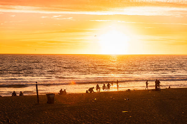 People stroll toward the shoreline as the sun sets over Santa Monica Beach, creating silhouettes and glowing sand at the iconic pier entrance.