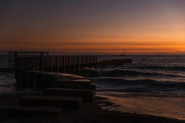 A peaceful sunset scene featuring a lone sailboat drifting on the horizon under a vivid orange and amber sky. The golden reflection stretches across the calm waves and smooth shoreline, creating a tranquil, cinematic moment that evokes solitude, freedom, and serenity. This minimalist coastal image is ideal for travel, mindfulness, marine lifestyle, or peaceful escape concepts.
