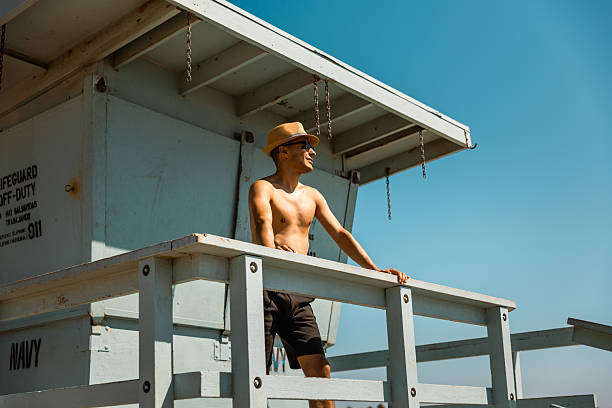 Portrait of a shirtless man wearing a straw hat and sunglasses standing on a pastel-blue lifeguard tower at Venice Beach, Los Angeles. He gazes into the distance with confidence under a bright blue sky. This sunny and vibrant summer image reflects freedom, relaxation, and the iconic California beach lifestyle with a stylish, laid-back attitude.
