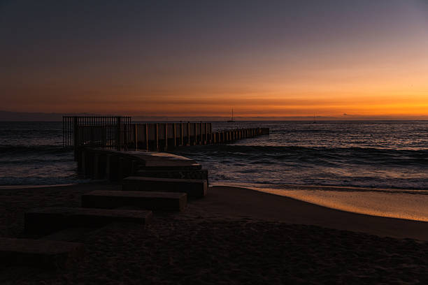 A peaceful sunset scene featuring a lone sailboat drifting on the horizon under a vivid orange and amber sky. The golden reflection stretches across the calm waves and smooth shoreline, creating a tranquil, cinematic moment that evokes solitude, freedom, and serenity. This minimalist coastal image is ideal for travel, mindfulness, marine lifestyle, or peaceful escape concepts.