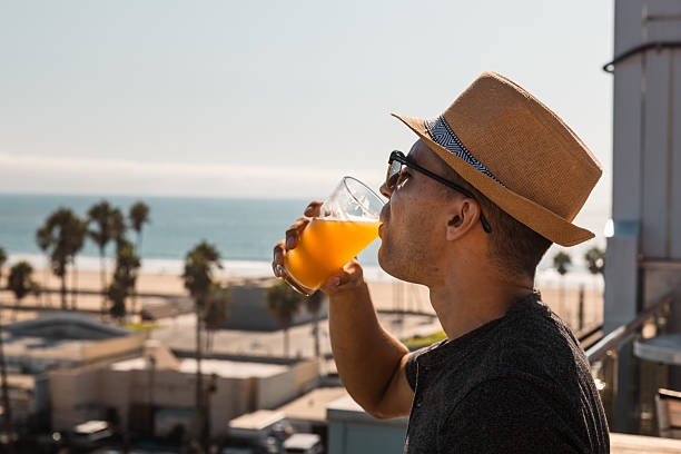 A fashionable man in sunglasses and a straw fedora hat is captured mid-sip of a refreshing drink on a sunny rooftop terrace in Venice Beach, Los Angeles. The Pacific Ocean and palm trees provide a dreamy coastal backdrop, highlighting themes of travel, relaxation, modern lifestyle, and summer leisure in Southern California.