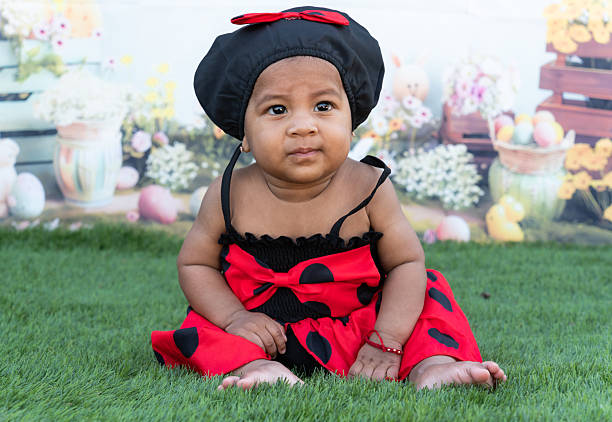 Cute baby girl wearing a ladybug costume sits on the grass in front of an easter-themed backdrop