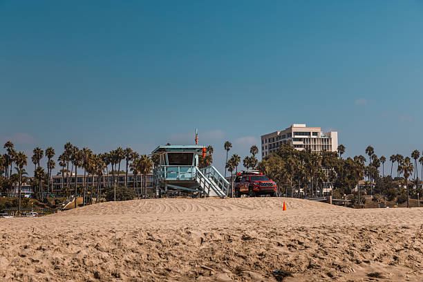 A bright red lifeguard rescue vehicle is parked beside a light blue lifeguard tower atop a sandy hill at Venice Beach in Los Angeles, California. Palm trees, a modern hotel, and blue skies create a distinctive coastal urban backdrop. The scene captures the iconic Californian beach safety infrastructure under the midday sun.