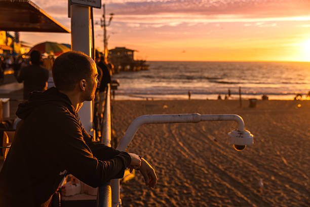 A man stands quietly at the edge of the Santa Monica Pier, gazing toward the horizon as the sun sets over the Pacific Ocean. The warm, golden-hour light paints the sand and waves in rich amber tones, creating a cinematic mood. Crowds fill the background in soft blur, while the foreground captures a reflective moment of solitude and wonder in California’s iconic coastal setting.