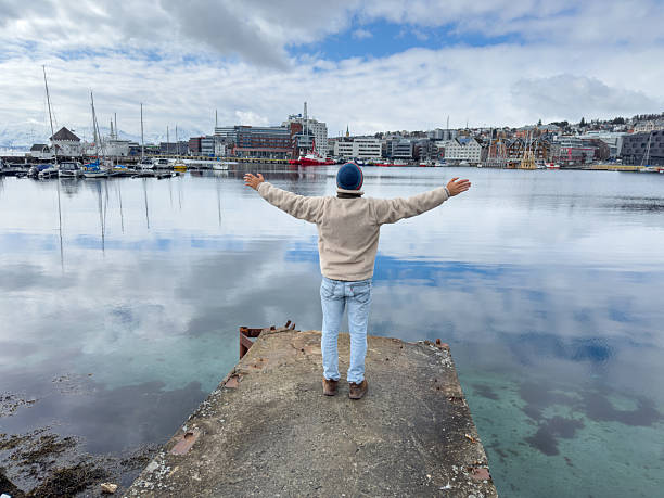 Individual standing on pier with arms wide open, overlooking serene harbor waters. Peaceful urban landscape with boats and waterfront buildings. Ideal for travel and inspiration themes.