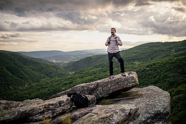 A young bearded man, dressed in relaxed hiking clothes and sturdy boots, stands proudly on a rock at the top of a hill, his backpack still on his shoulders. After a challenging hike, he takes in the breathtaking view around him — surrounded by rich greenery, with dramatic clouds above and sunlight breaking through.