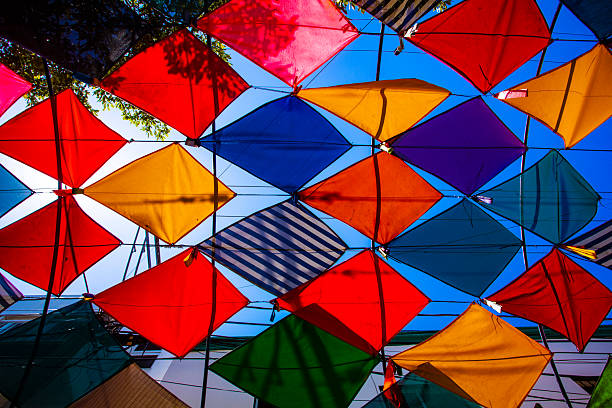 Brightly colored kites hanging as decorations above a narrow street, viewpoint looking straight up, blue sky and bright sunlight