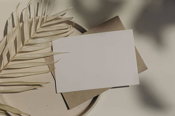istock Blank horizontal greeting card, invitation mockup, craft envelope in sunlight, beige table background. Floral shadows overlay. Palm leaf on plate. Neutral web banner. Elegant stationery. Flat lay. Top view. 2219695549