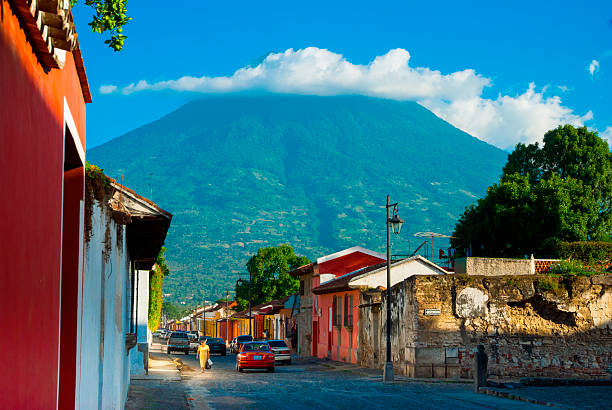 Street scene in Antigua Guatemala with colorful colonial homes, locals, parked vehicles and a cloud covered volcano in the background. Vibrant culture, history, and majestic natural beauty merge.