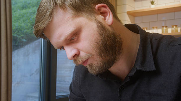 istock Bearded man in a dark shirt looks down with intense concentration, likely working or reading near a window in natural light. 2219704023