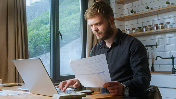 istock Focused bearded man sits by a window, reviewing printed documents and typing on a laptop, suggesting work or financial analysis. 2219704391