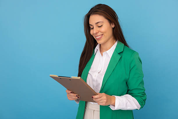 istock Confident professional woman reviewing documents, smiling brightly with clipboard in hand against vivid blue backdrop 2219706033