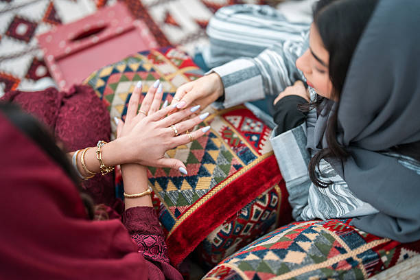 Two Arabic young adult females sit comfortably in a traditional Arabian Majlis, surrounded by vibrant textiles. One wears a hijab, and the other is engaged in a cultural exchange, highlighting rich heritage.