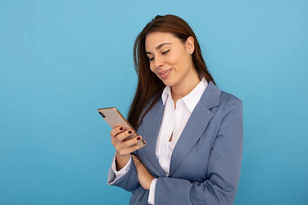 istock Smiling young businesswoman using a smartphone while standing against a vibrant blue backdrop, exuding positivity and professionalism 2219708005