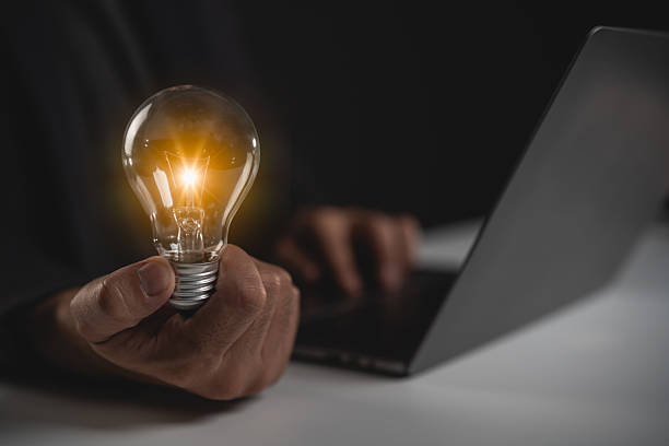 istock Light bulb held by hand showcasing innovation and creativity in the digital workspace with laptop on table in dark environment 2219727860