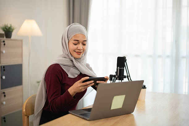 Young woman with hijab engaging in podcast recording while using mobile phone at modern workspace, surrounded by soft lighting and natural decor