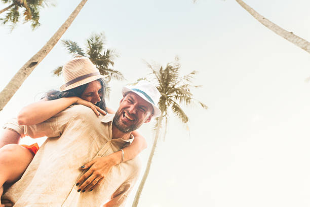 Laughing couple in summer hats enjoy piggyback moment under palm trees in tropical setting with warm golden light and carefree energy. People travel enjoy honeymoon outdoors