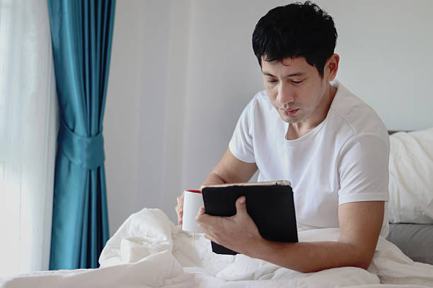 istock Asian man using tablet while drinking coffee in bed after waking up in morning in bedroom. Man working on his tablet during lying down on comfortable bed. Sleepy man surfing internet in bed at home. 2219749481