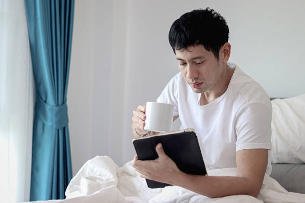 istock Asian man using tablet while drinking coffee in bed after waking up in morning in bedroom. Man working on his tablet during lying down on comfortable bed. Sleepy man surfing internet in bed at home. 2219749484