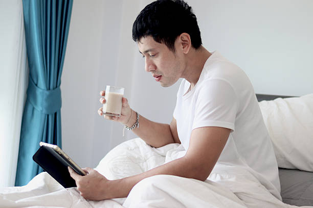 istock Asian man using tablet while drinking almond milk in bed after waking up in the morning in bedroom. Man working on his tablet during lying down on comfortable bed. Sleepy man surfing internet in bed. 2219749490