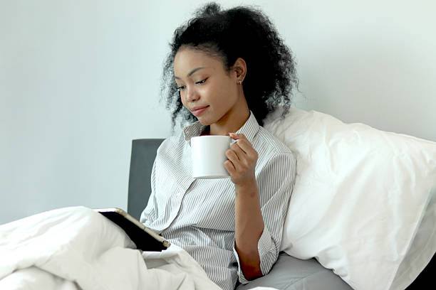 istock Beautiful African woman with curly hair drinking coffee while using tablet after waking up in morning in bedroom. African female holds coffee mug, works on tablet during lie down on comfortable bed. 2219749711