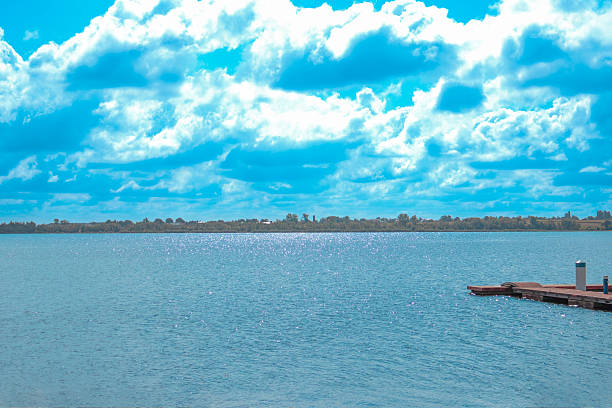 Bright blue sky with scattered clouds reflects on calm water near a dock.