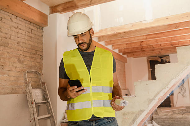 istock Male Contractor in safety vest and helmet using smartphone holding blueprints on construction site 2219763481