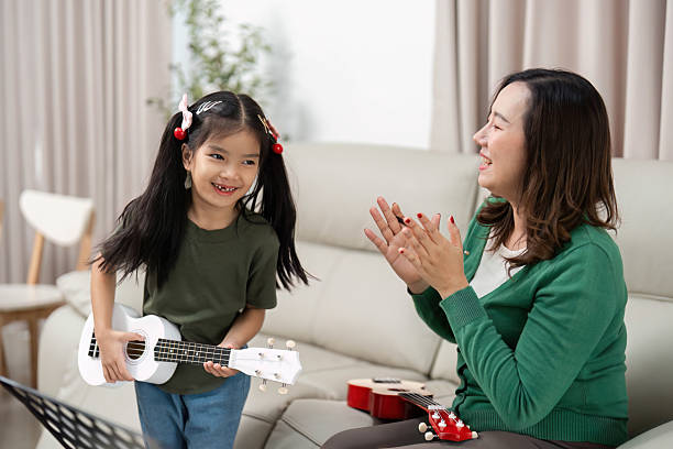 istock Learning and Fun. Young girl joyfully playing ukulele while mother claps in encouragement. 2219769776