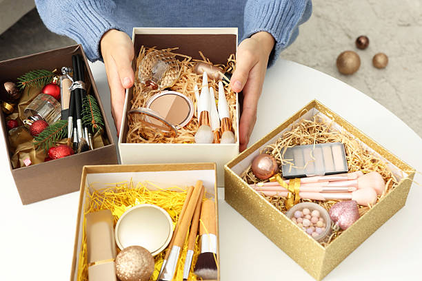 istock Woman holding Christmas gift boxes with makeup brushes and cosmetics at white table, closeup 2219770038