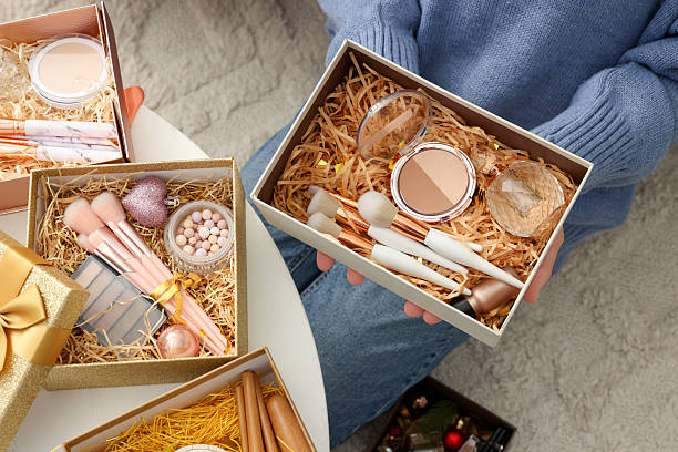 istock Woman holding Christmas gift boxes with makeup brushes and cosmetics at white table, above view 2219770101