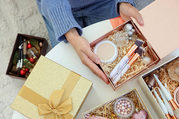 istock Woman holding Christmas gift boxes with makeup brushes and cosmetics at white table, above view 2219770114