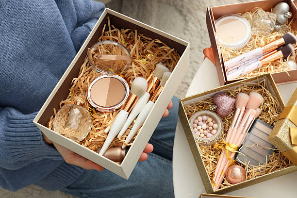 istock Woman holding Christmas gift boxes with makeup brushes and cosmetics at white table, closeup 2219770116