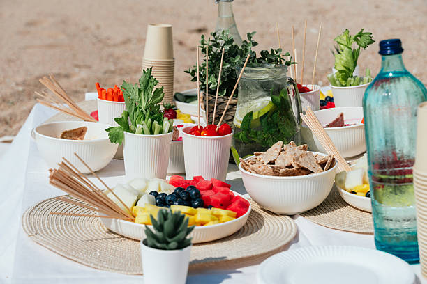 Fresh fruit, vegetables, and snacks are arranged on a table set up on a sandy beach, ready for a healthy vegan meal