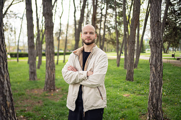 A young bald man with a light jacket stands confidently among tall trees in a park.