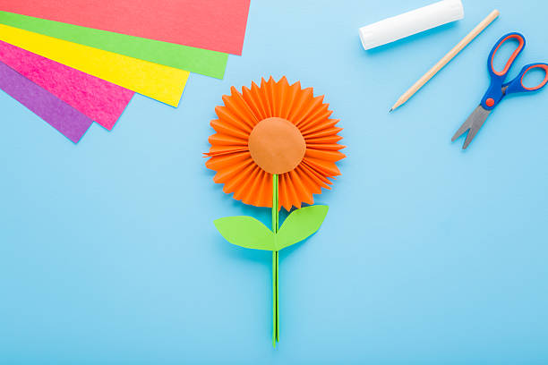 istock Orange flower, scissors, glue stick, pencil and colorful application paper on light blue table background. Pastel color. Closeup. Creating festive decoration. Top down view. 2219786068