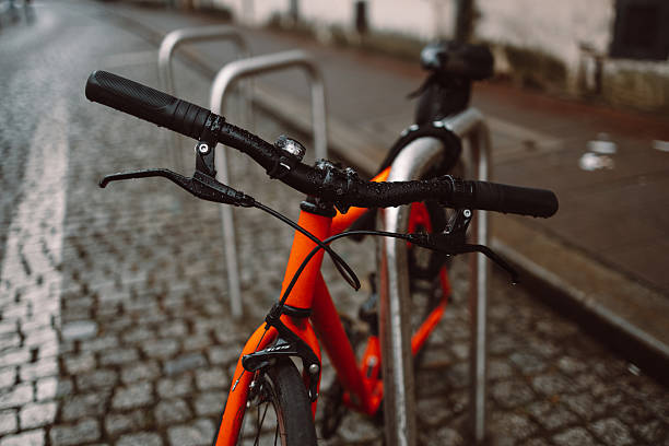 Close-up of an orange bicycle's handlebars, locked to a metal bike rack on a cobblestone street. The wet pavement and blurred background evoke a quiet, rainy day in the city.