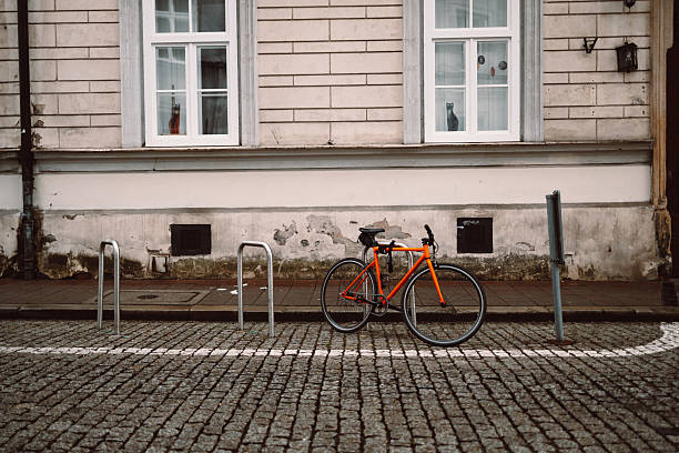Bright orange bicycle is parked and secured to a metal bike rack on a cobblestone street. Behind it stands a historic beige building with white-framed windows, evoking a calm, urban European setting