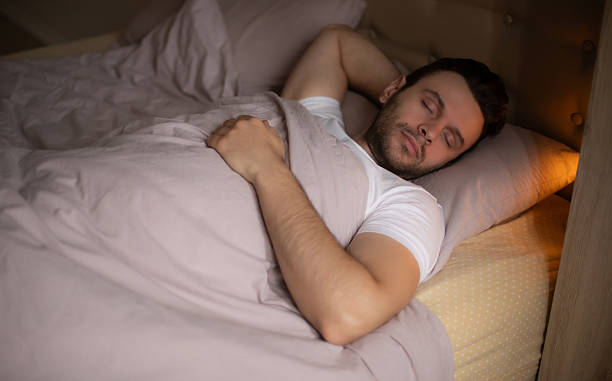 istock Young man sleeping peacefully in bed under soft bedding, wearing a white T-shirt, resting on a pillow in a cozy, warmly lit bedroom, capturing calm and relaxation. 2219795268