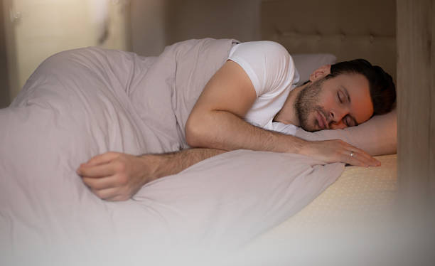 istock Young man peacefully sleeping on his side in bed, wearing a white T-shirt, wrapped in soft blankets, in a calm, cozy bedroom with soft morning light creating a serene atmosphere. 2219795292
