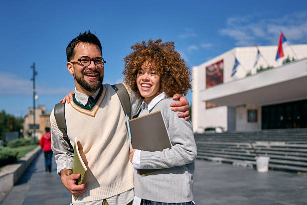 Two cheerful multi-ethnic students are walking embraced outside their university building, holding books and notebooks, smiling and looking away