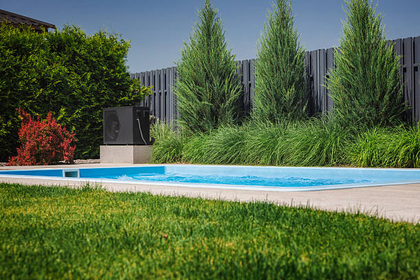 istock Modern heat pump installed next to a backyard pool, used for heating water, surrounded by green lawn, conifers, and privacy fence in a landscaped garden. 2219801610